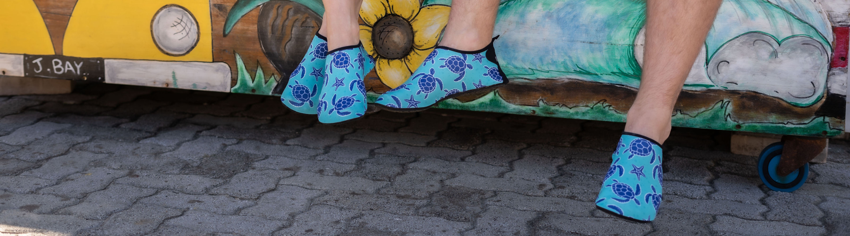Close-up of two people wearing matching Aqua Turtle Kaalfööt kicks with sea turtle designs, sitting on a colourful painted bench in J-Bay.